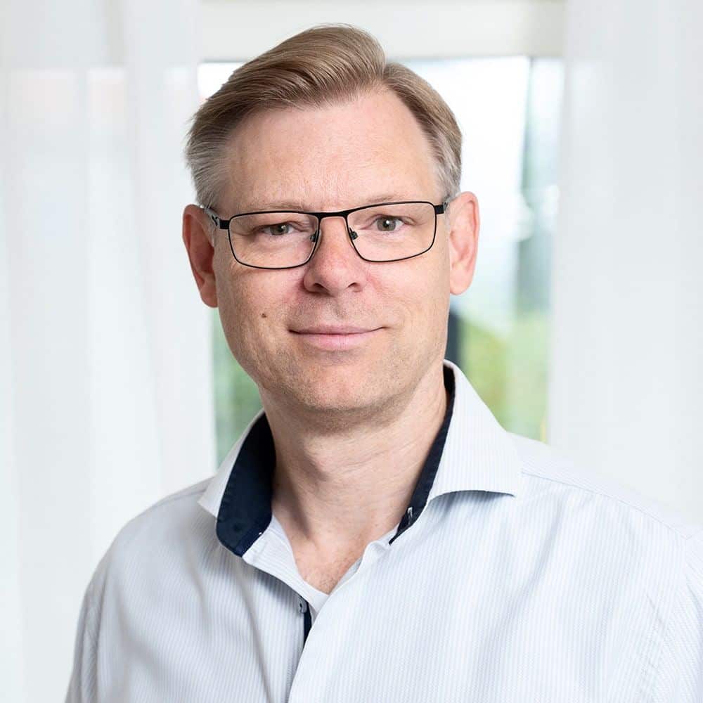 En man med kort ljust hår och glasögon har på sig en ljus skjorta och står framför vita gardiner med ett neutralt ansiktsuttryck.