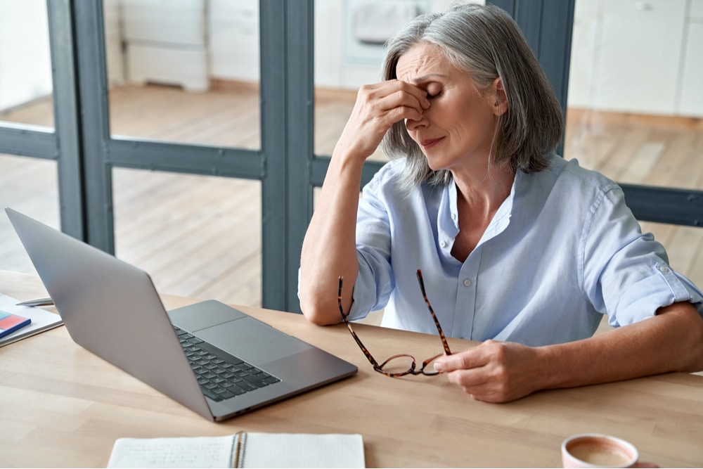 Äldre kvinna sitter vid ett bord framför en laptop, håller glasögonen i ena handen och gnuggar sig i ögonen med den andra, ser trött eller stressad ut. Hon kan ha nytta av svensk personal som erbjuder trygghet och stöd.